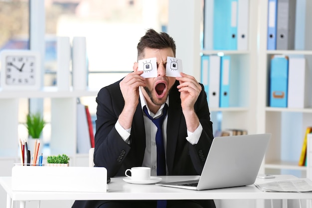 Young business man with fake eyes painted on paper stickers yawning at workplace in office