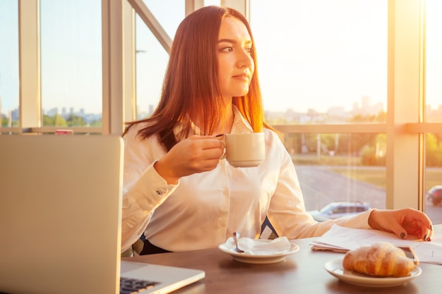 Young business woman drinks coffee and works behind laptop with papers.