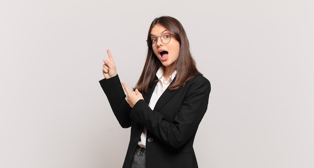 Young business woman feeling joyful and surprised, smiling with a shocked expression and pointing to the side