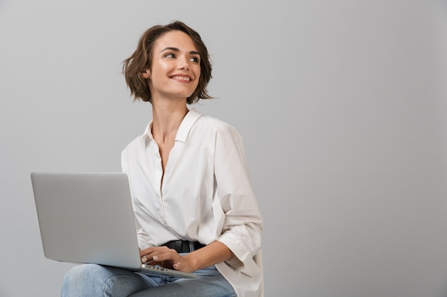 Young business woman posing isolated over grey wall sitting on stool using laptop computer