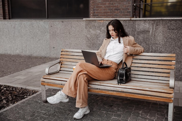 Young business woman sitting on a bench with a laptop of the modern office building background