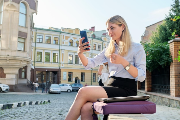 Young business woman sitting outdoors talking video online using smartphone