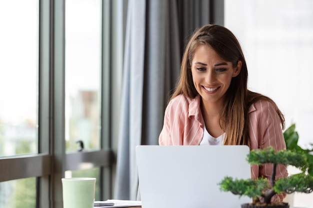 Young business woman using laptop at home looking at screen chatting reading or writing email sitting on couch female student doing homework working on research project online