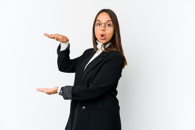 Young business woman on white wall shocked and amazed holding a blank space between hands.