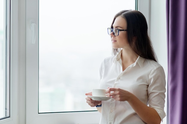 Young business woman with cup of coffee near window in office, city in window for autumn winter season. Coffee break, positive woman teacher, psychologist, consultant, secretary