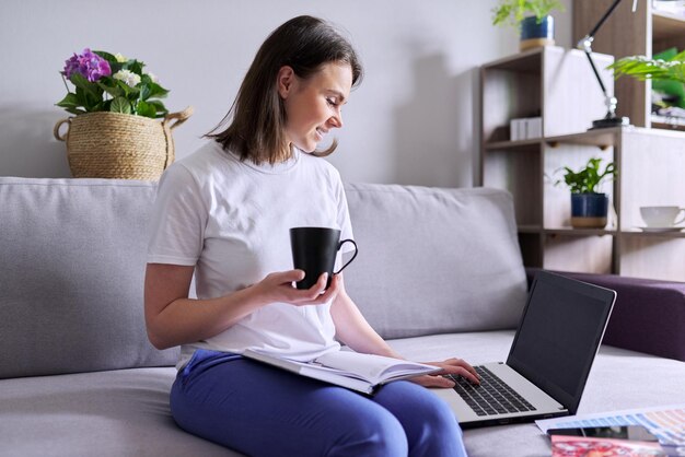 Young business woman with laptop computer, cup of coffee sitting on couch, home interior background. Remote employment, freelance, online education and teaching, career lessons and development concept