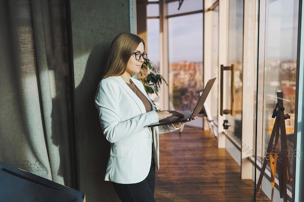 A young business woman with long blond hair in a white jacket and glasses stands with a laptop in her hands in an office space Work in a modern office with large windows