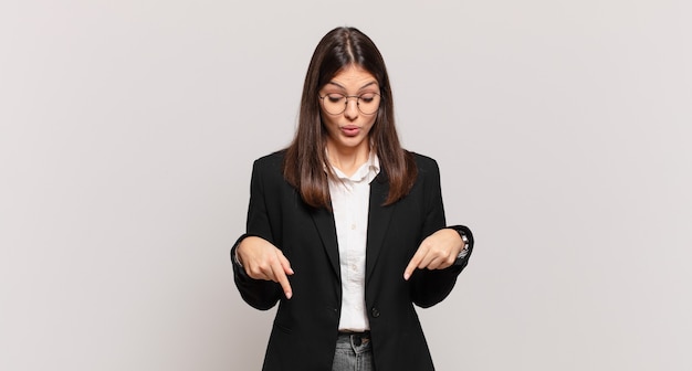 Young business woman with open mouth pointing downwards with both hands, looking shocked, amazed and surprised