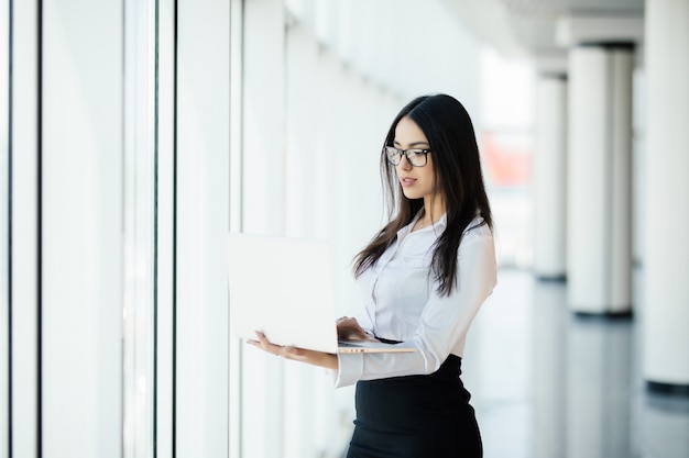 Young business woman working in her luxurious office holding a laptop standing against panoramic window with a view on business district