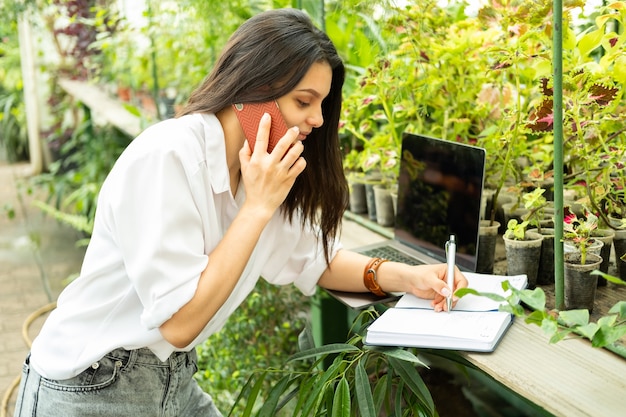 Young business women gardener talking on phone, writing in notebook in greenhouse.