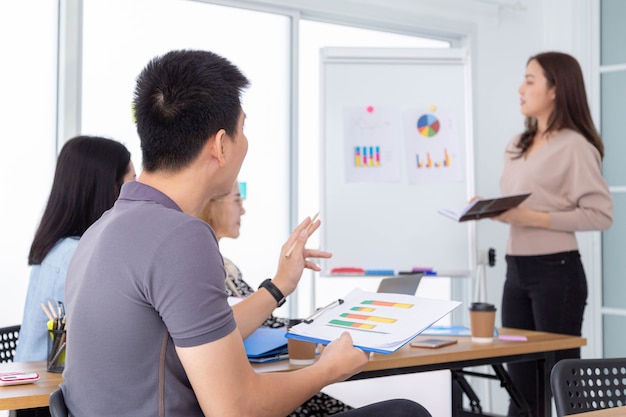 A young businessman asks for information from the young businesswoman presented in front of the conference room.