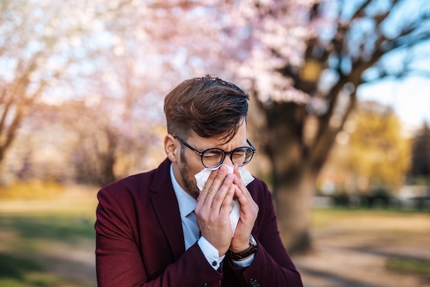 Young businessman blowing his nose in park. Allergy, flu, virus concept.