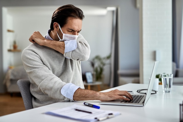 Young businessman coughing into elbow while wearing protective face mask and working on a computer at home