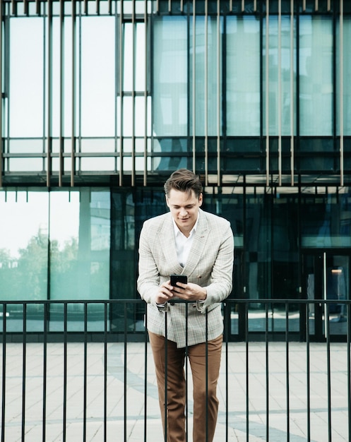 Young businessman dressed in formal clothes standing outside a glass building and using mobile phone