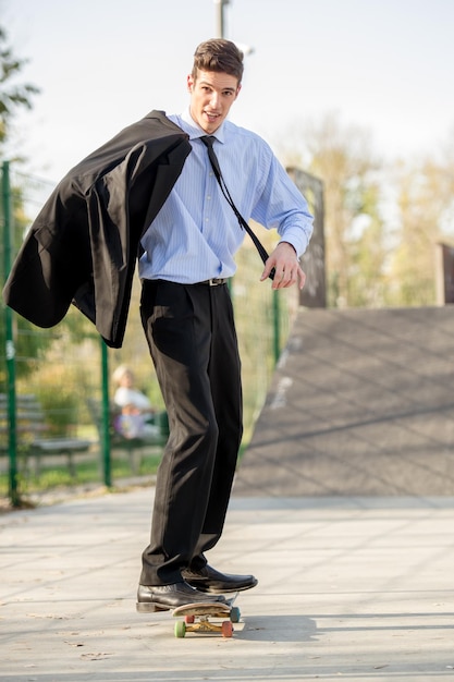 Young businessman, elegantly dressed, holding a jacket draped over shoulder standing on a skateboard. Looking at camera