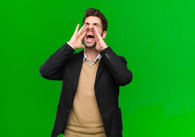 Young businessman feeling happy, excited and positive, giving a big shout out with hands next to mouth, calling out on green