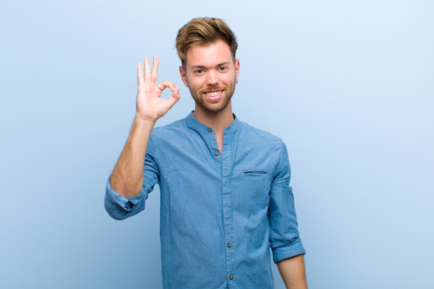 Young businessman feeling happy, relaxed and satisfied, showing approval with okay gesture, smiling against blue wall