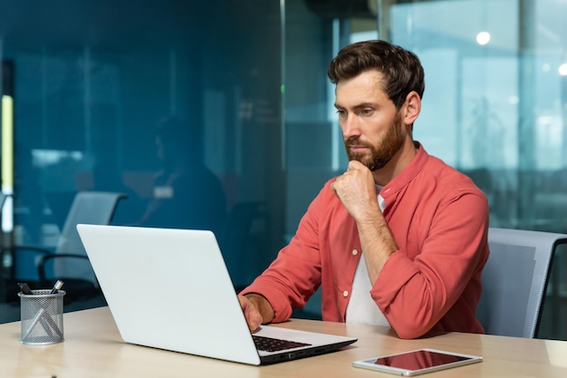 A young businessman freelancer designer in a red shirt sits in the office at the table and works on