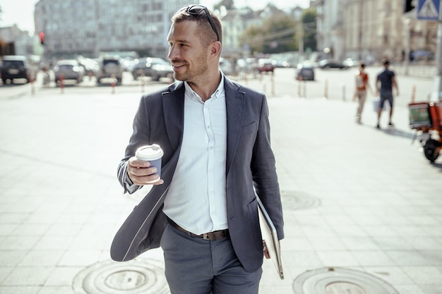 Young businessman holding his laptop while drinking coffee. Standing on the street.