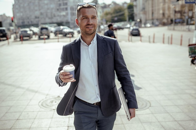 Young businessman holding his laptop while drinking coffee. Walking on the street.