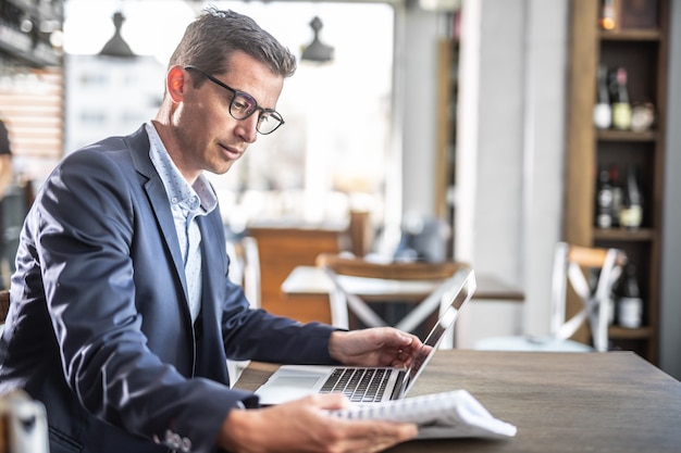 Young businessman in a jacket and glasses reads in a cafeteria with a computer on a table in front of him.