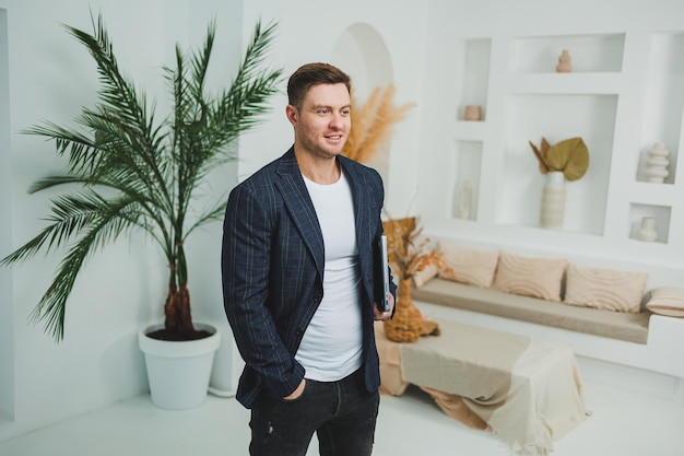 A young businessman in a jacket with a laptop stands in a bright room and works remotely Work from home A freelancer works online on a laptop