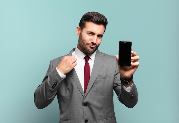 Young businessman looking arrogant, successful, positive and proud, pointing to self and showing his phone screen