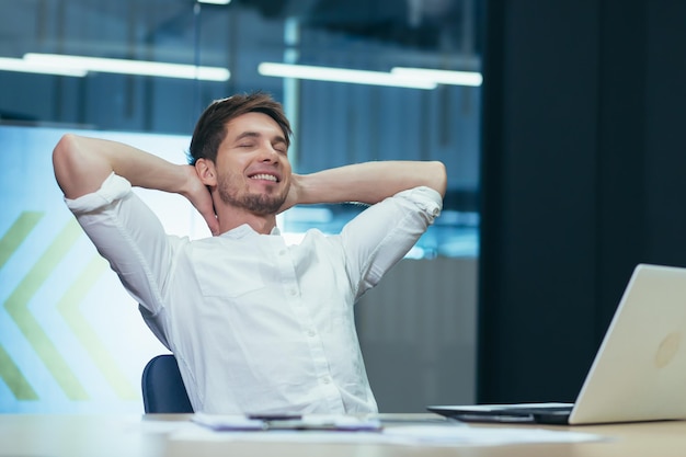 A young businessman in the office is resting and dreaming throwing his hands behind his head a man at work working with a laptop