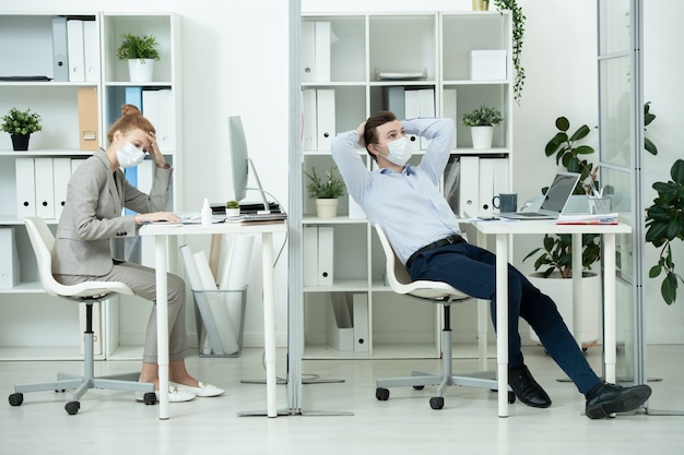 Young businessman in protective mask brainstorming in front of laptop while his female colleague thinking during work over new project