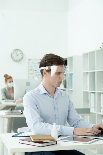 Young businessman in protective screen on head and formalwear looking at laptop display while working with data against female colleague