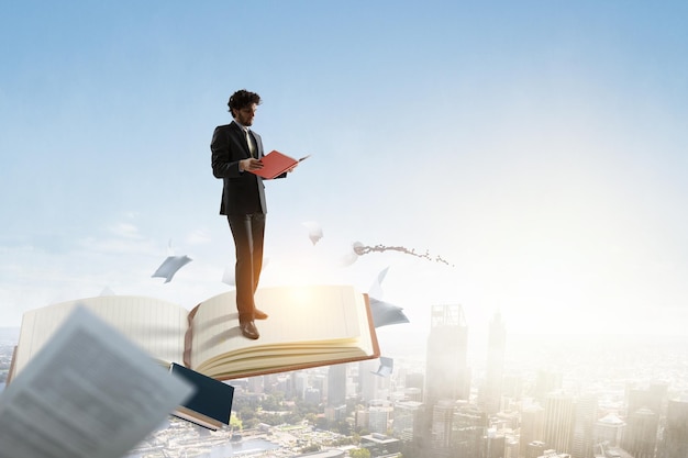 Young businessman reading a book standing on a big notebook. Mixed media