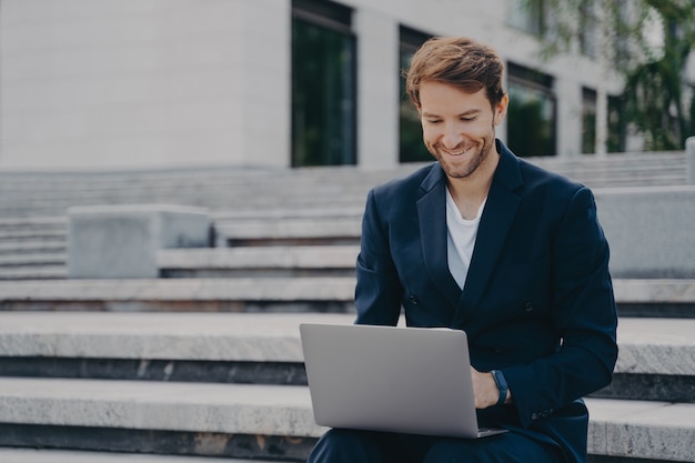 Young businessman sits on steps uses laptop organizes meeting online with investors