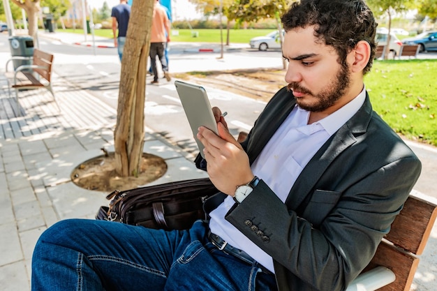 Young businessman sitting on a bench in the park holding his tablet