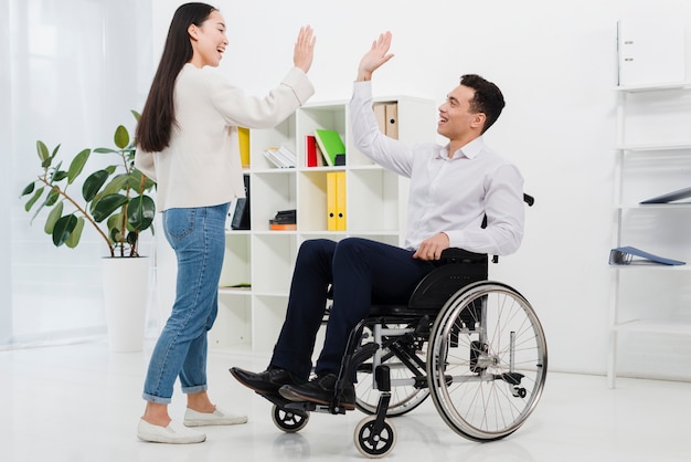 Young businessman sitting on wheelchair giving high-five to her female colleague in the office