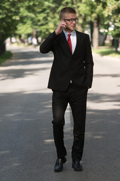 Young Businessman Talking On The Phone While Walking Outside In Park