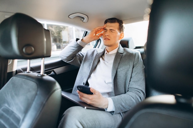 Young businessman tired from work holding hand to his head while sitting in the back seat of the car