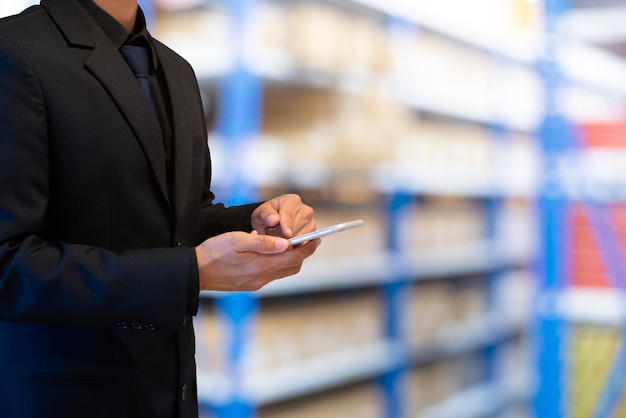 A young businessman wearing a black suit is using a tablet and the Internet to check stocks in a warehouse in preparation for delivery to customers