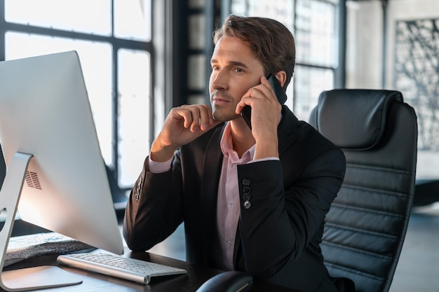 Young businessman wearing black suit sitting to table with computer making a call Office manager with phone working in modern office with glass windows blurred background concept of work