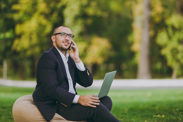 Young businessman in white shirt, classic suit, glasses. Man sit on soft pouf, talk on phone, work on laptop pc computer in city park on green lawn outdoors on nature. Mobile Office, business concept.