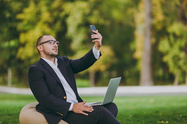 Young businessman in white shirt, classic suit, glasses. Man sit on soft pouf with laptop pc computer, doing selfie on mobile phone in city park on green lawn outdoors. Mobile Office business concept.