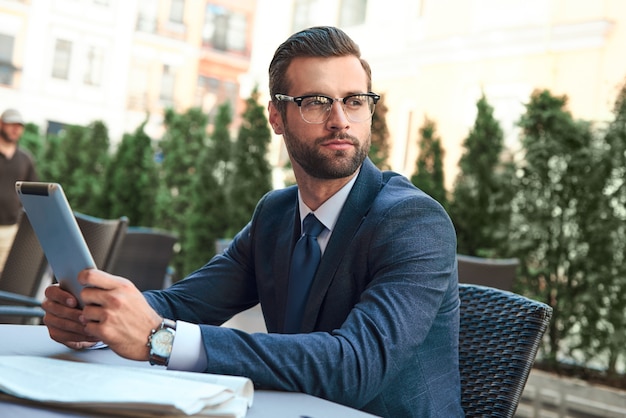 Young businessman with a beard and wearing glasses is sitting and looking at the tablet