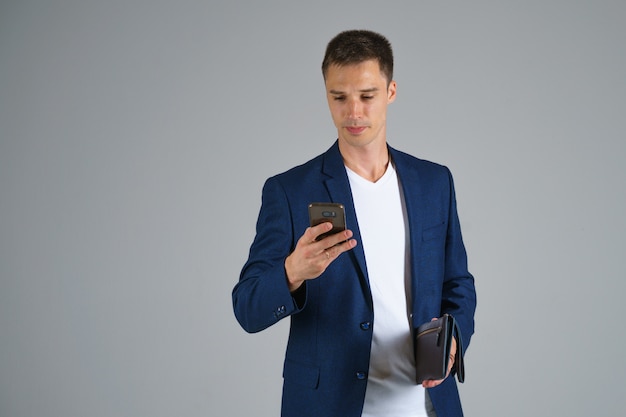 A young businessman with short hair in a blue jacket and white t shirt is holding a phone in his hand and talking against a gray background