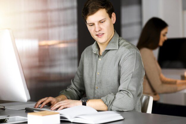 Young businessman working on his computer sitting at the desk in office. Headshot portrait of a man.