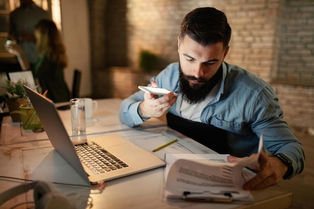 Young businessman working late on paperwork and talking over smart phone's speaker in the office