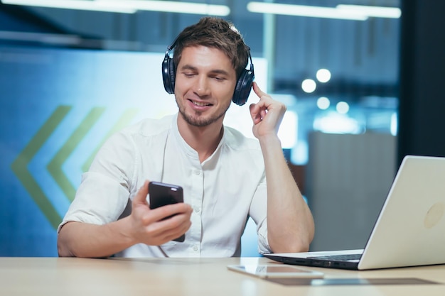 Young businessman works in the office on a laptop listens to music in big headphones uses the application to listen to online books and podcasts uses the phone