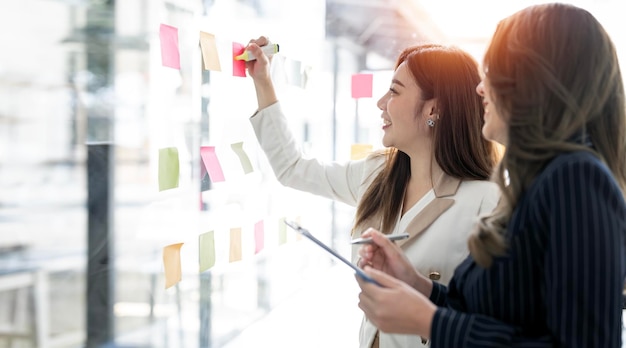 Young businesswoman creative team using post it notes in glass wall to writing strategy business plan to development grow to success