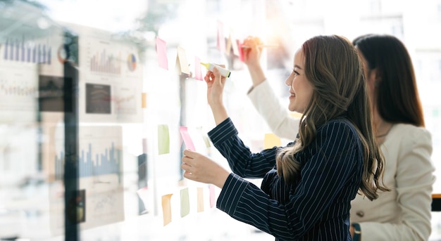 Young businesswoman creative team using post it notes in glass wall to writing strategy business plan to development grow to success