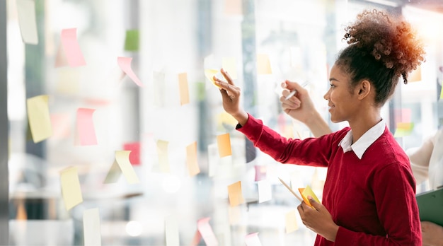 Young businesswoman creative team using sticky notes in glass wall to writing strategy business plan to development grow to success