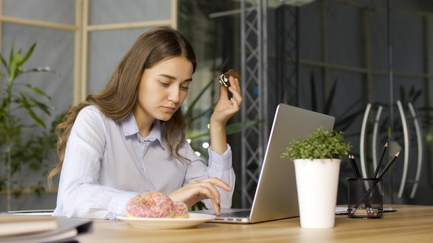 Young businesswoman eating a glazed donut while working on laptop in office