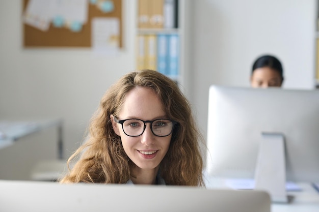 Young businesswoman in eyeglasses sitting at table in front of monitor and working on computer